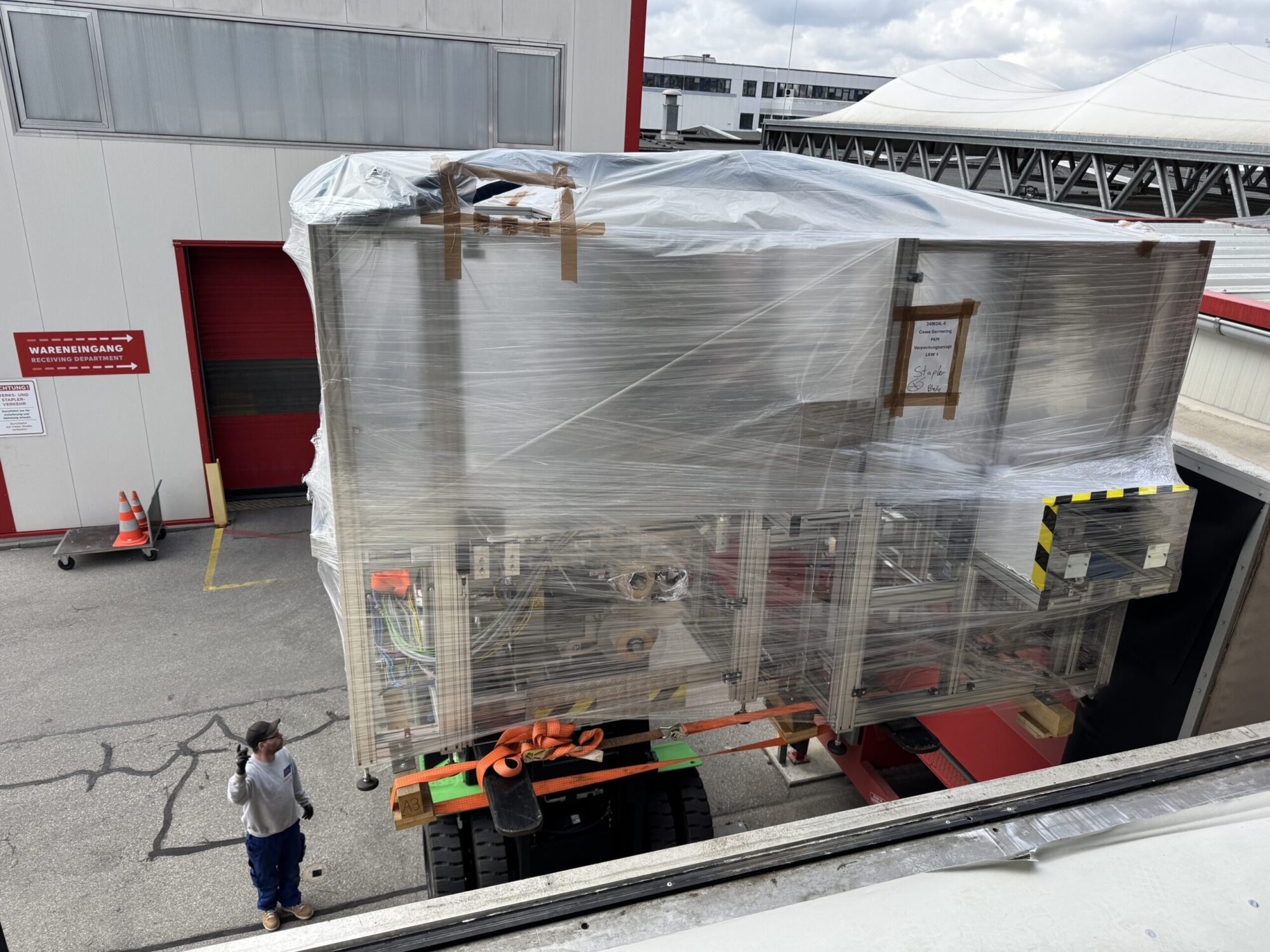 Ein großes, in Folie verpacktes Objekt auf einem Lkw-Anhänger vor einem Gebäude, daneben zwei Personen.