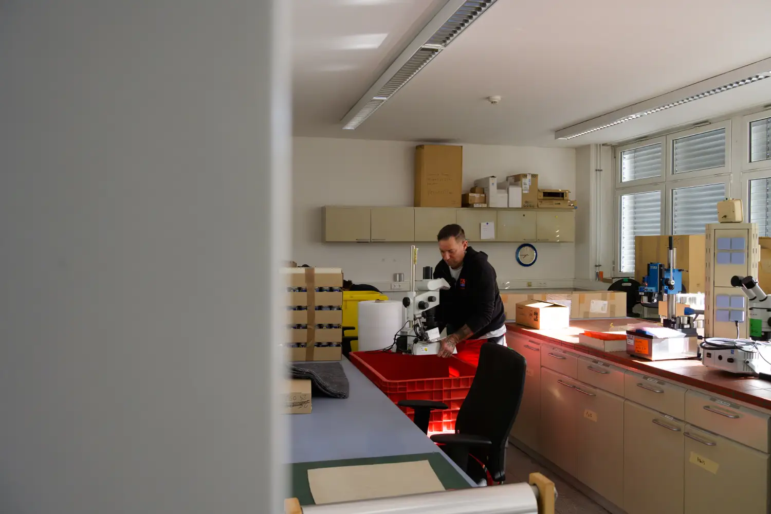 Person arbeitet mit einem Mikroskop in einem Labor mit Schränken und Regalen.