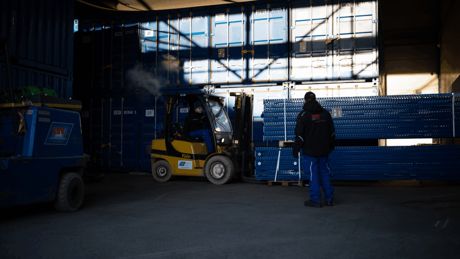 Gabelstaplerfahrer lädt schwere Metallregale vor Containern, ein weiterer Mitarbeiter koordiniert die Ladung.