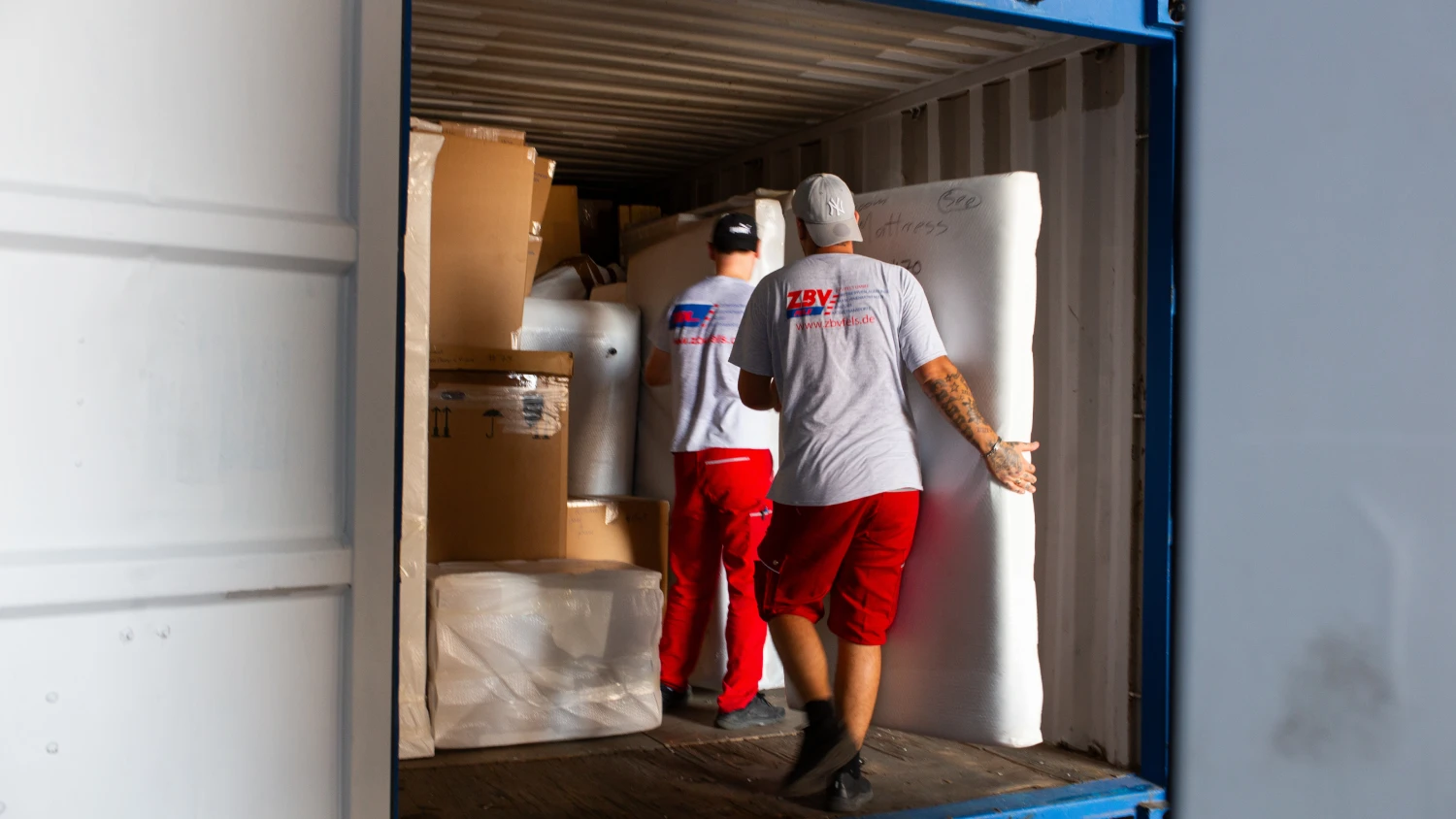 Zwei Mitarbeiter von ZBV Fels verladen gepolsterte Möbel und Kartons in einen Lagercontainer.