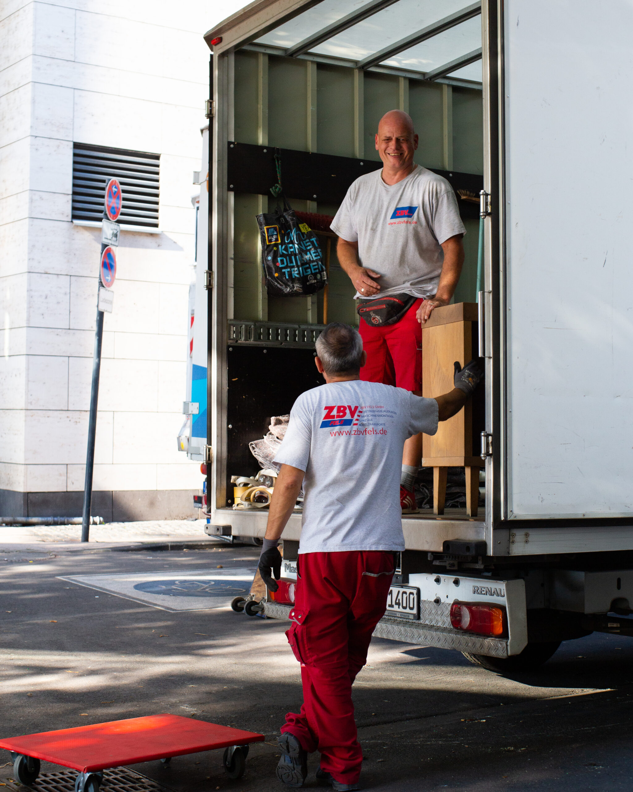 Zwei Mitarbeiter von ZBV Fels laden Umzugskartons aus einem Lieferwagen auf der Straße aus.