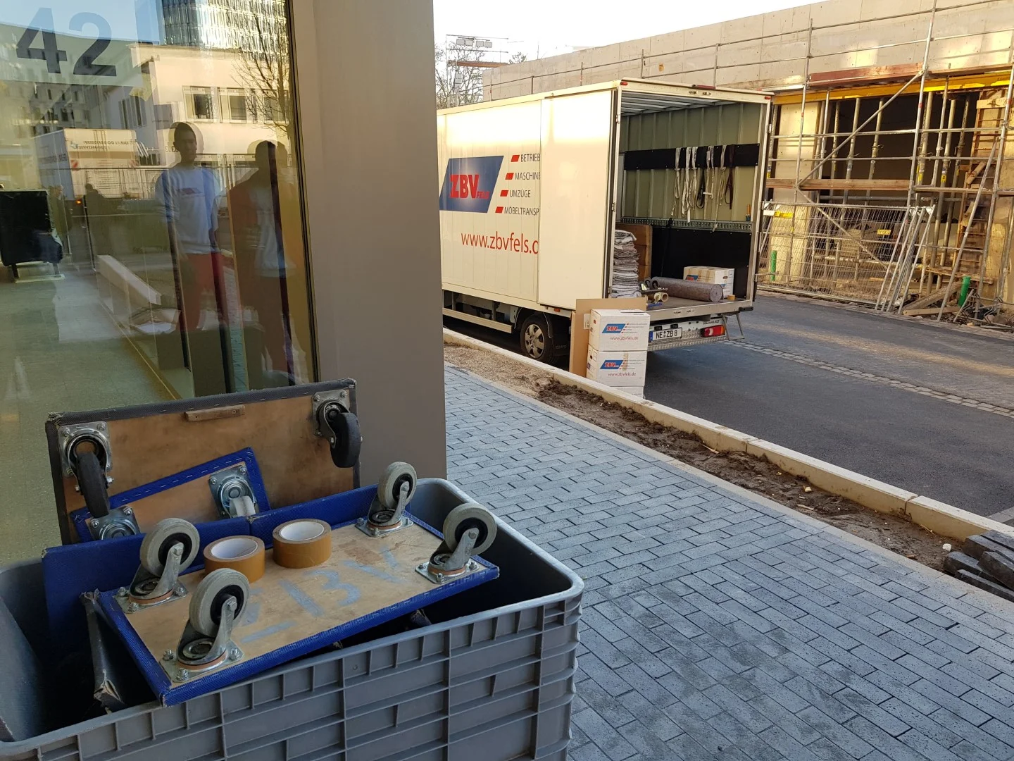 Lkw liefert Material an eine Baustelle, im Vordergrund steht ein Behälter mit Rollen.