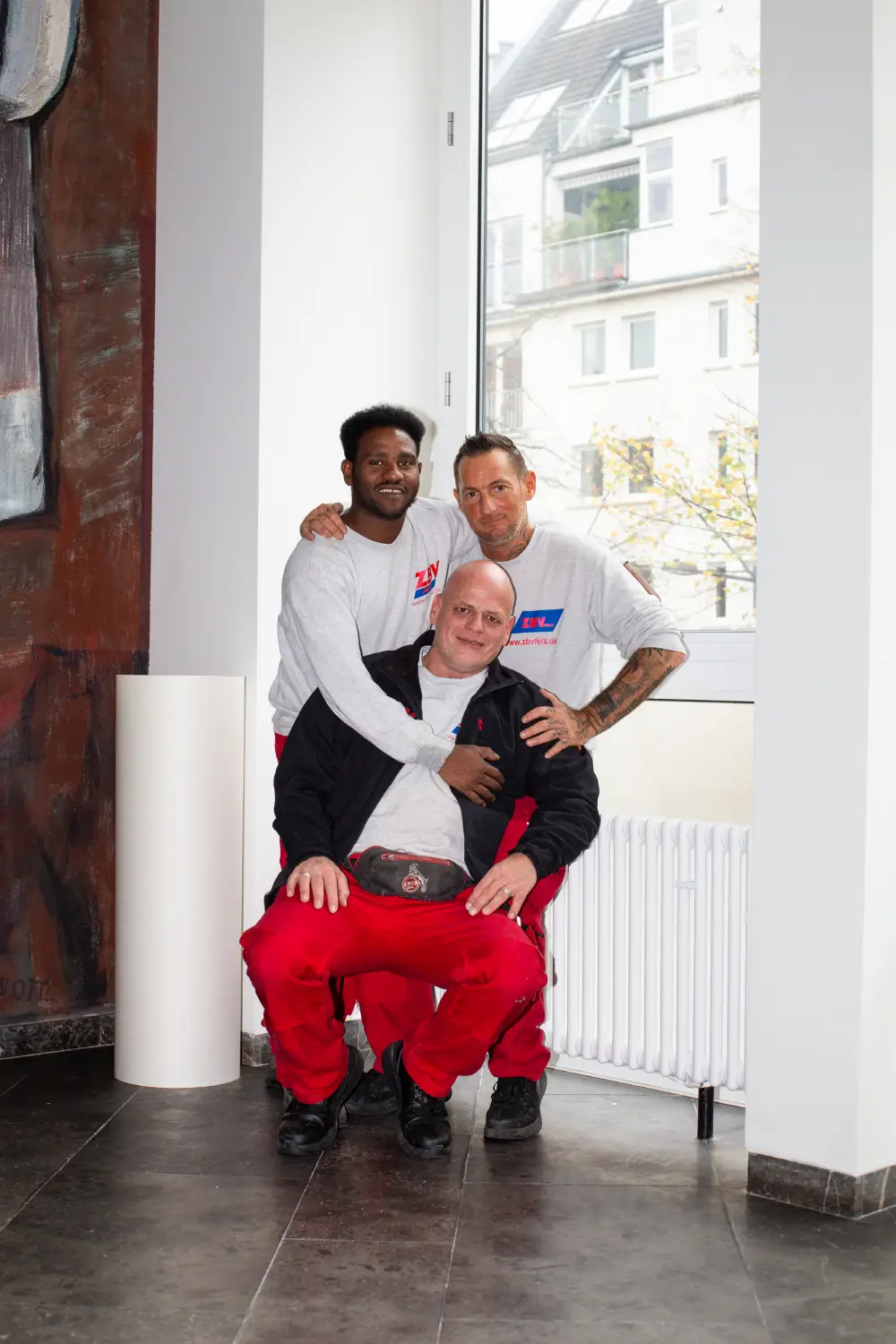 Drei Männer vom Team in einem hellen Raum, zwei stehen hinter einem sitzenden Mann, alle lächeln.
