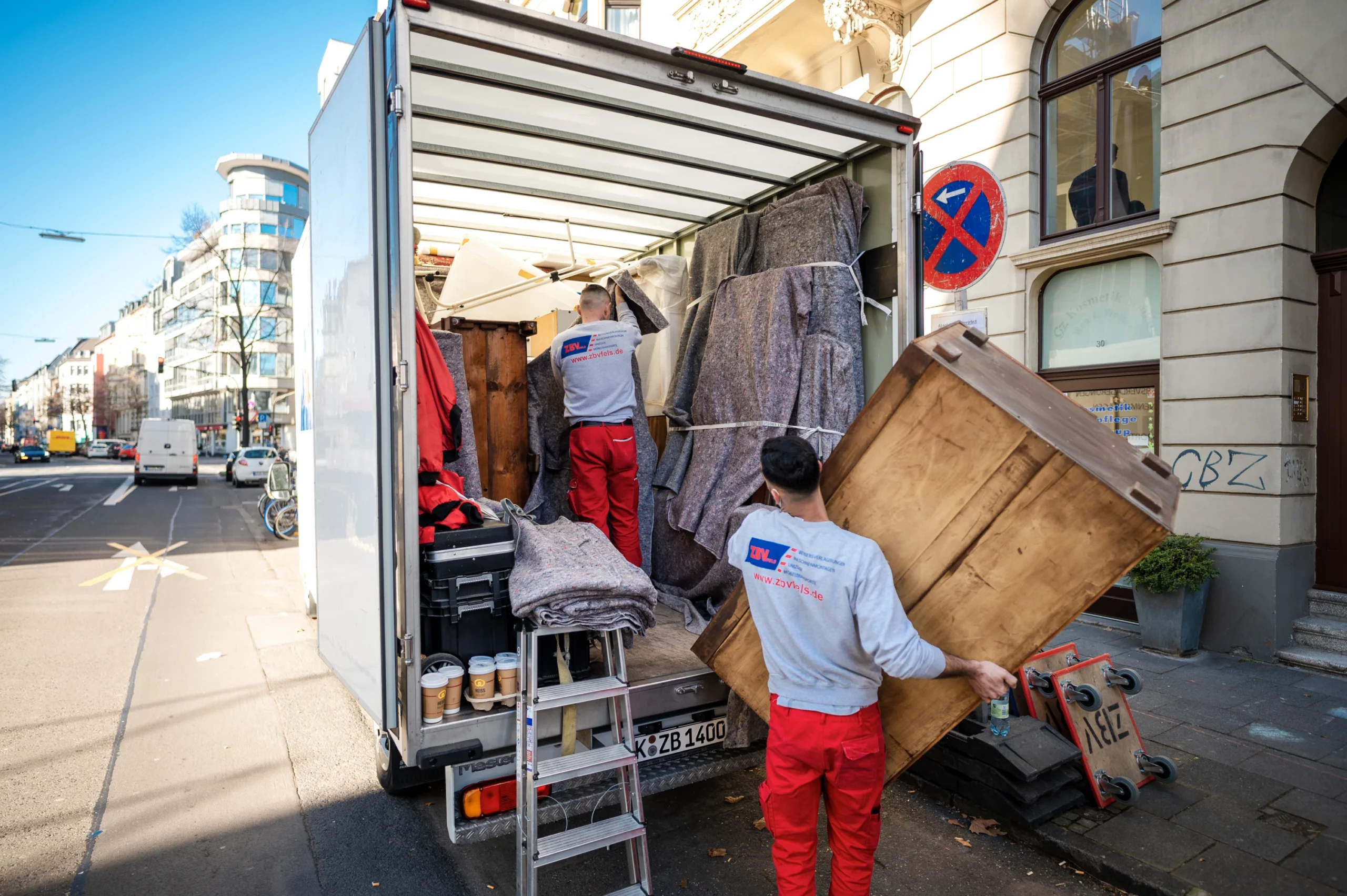 Zwei Umzugshelfer von ZBV Fels laden Möbel aus einem Lkw vor einem Wohnhaus aus.
