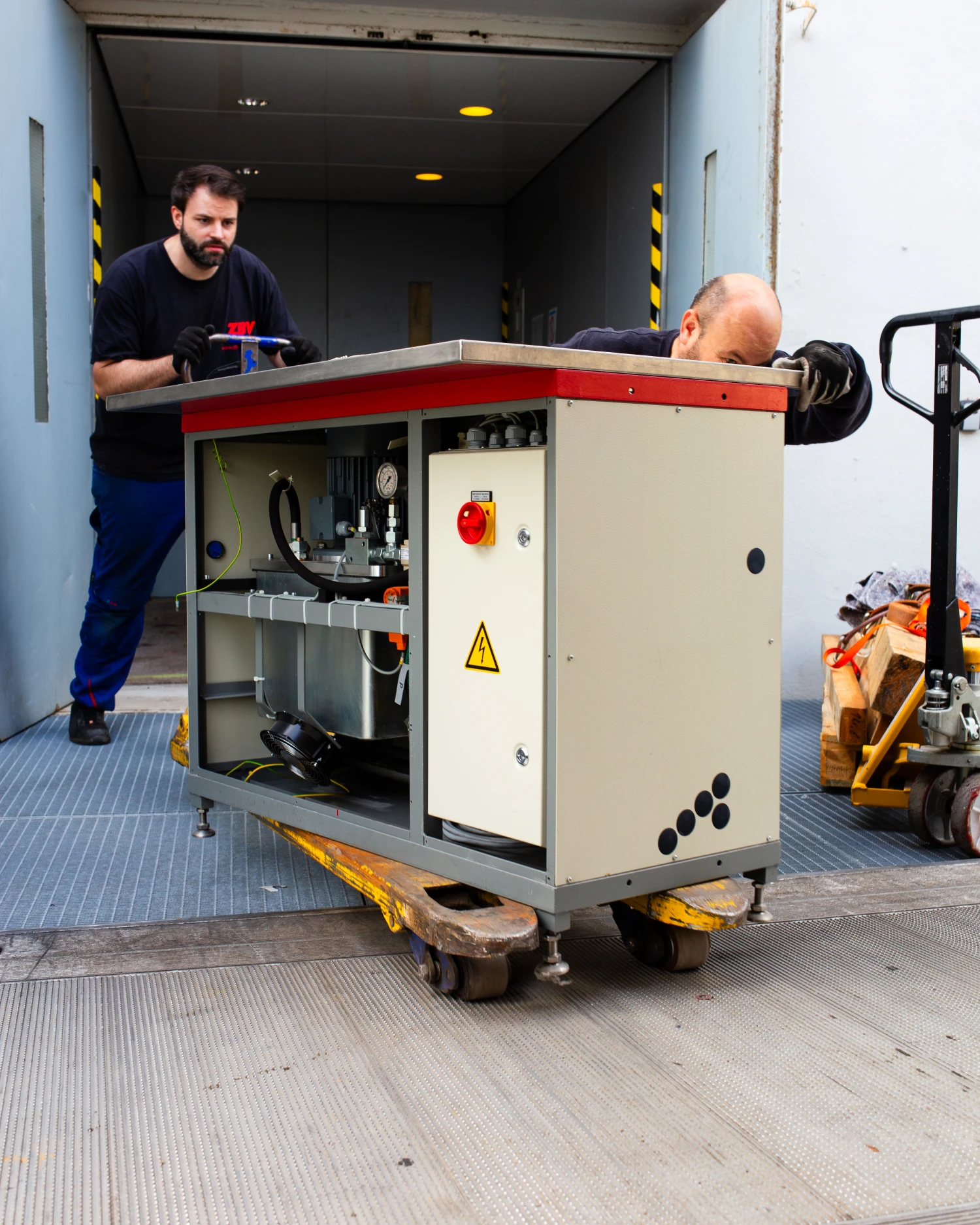 Zwei Männer bewegen eine große Maschine mit einem Hubwagen über eine Rampe.