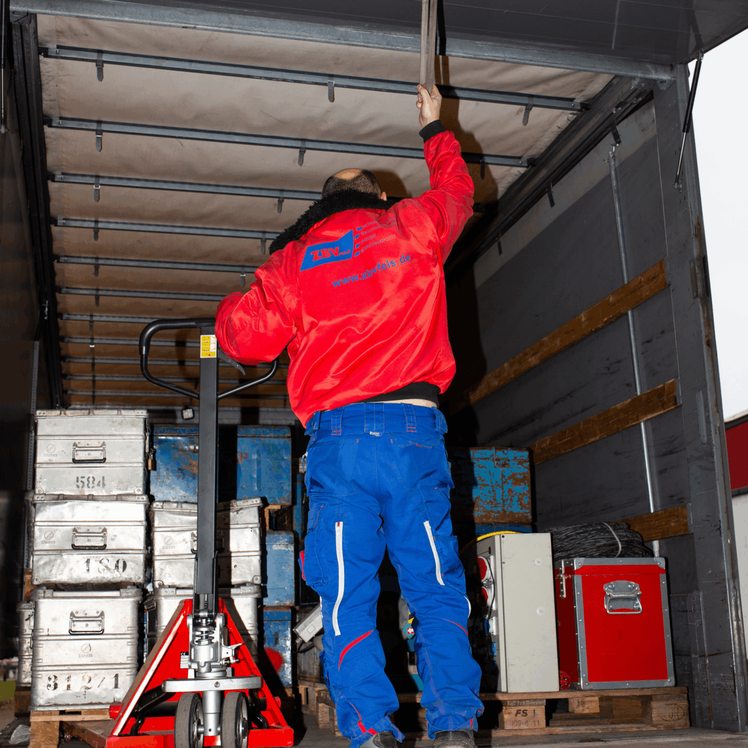 Mitarbeiter von ZBV Fels sichert Fracht im Lkw mit Hubwagen und stapelbaren Metallkisten.